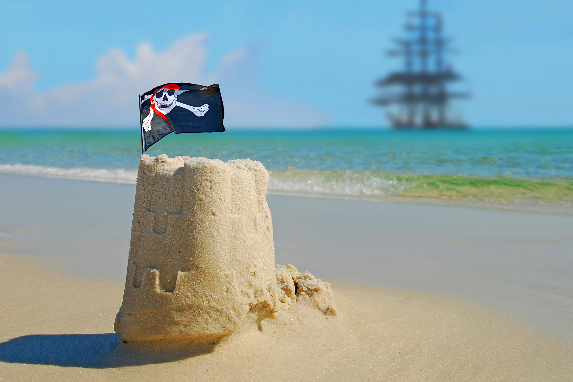 a small sandcastle on the beach with a pirate flag stuck in the top and in the distance a pirate ship in the water